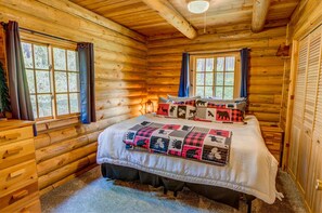 Ein Schlafzimmer in einer Blockhütte mit einem Bett, zwei Fenstern, einer Holzkommode und einer Steppdecke mit Tiermotiven.