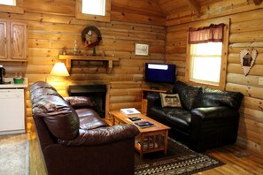 A cozy cabin interior with a fireplace, leather sofas, a television, and a small table. - Charming Cabin Rental in the Forest near Beckley, West Virginia (Hico)