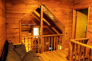 A cozy cabin interior with a lofted sleeping area, a wooden staircase, and a door leading to another room. - Charming Cabin Rental in the Forest near Beckley, West Virginia (Hico)