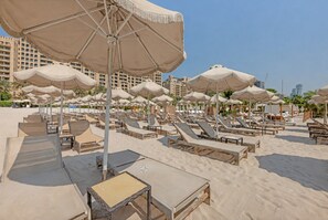 Ein Strand mit Reihen von Liegestühlen und großen Sonnenschirmen, manche mit Tischen, unter blauem Himmel.