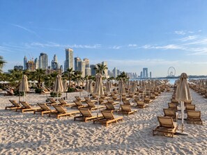 A beach with rows of sun loungers and umbrellas, a city skyline in the background, and palm trees. - Luxury One-Bedroom Seven Palm | Beach & Pool (Dubai)