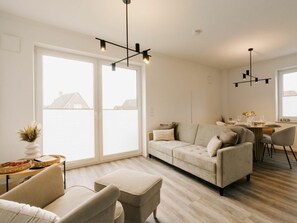 A modern living room with a sofa, ottoman, and dining area with chairs and a table. - Holiday Apartment in Greetsiel With Balcony (Krummhoern)