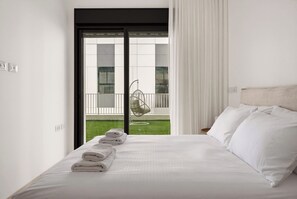 Ein Hotelzimmer mit Bett, weißen Leinen, Blick auf einen Balkon mit Hängesessel und einem ordentlich bezogenen Bett mit Handtüchern.