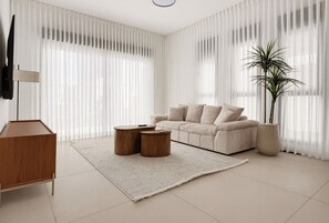Ein modernes Wohnzimmer mit einer Couch, zwei runden Holztischen, einer großen Topfpflanze und transparenten weißen Vorhängen.