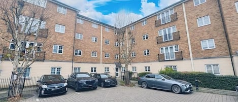 Un immeuble résidentiel à plusieurs étages, doté d’une façade en briques, d’une cour pavée et de plusieurs voitures garées.
