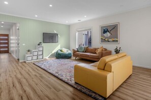 A living room with a yellow sofa, a blue bean bag, a patterned rug, a television, and a framed picture of a bird. - Beach & Bath - Torquay Family Getaway (Surf Coast)