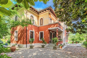 A two-story building with a red facade, multiple windows, and a garden with potted plants. - Vrbo Property (Mozzate)