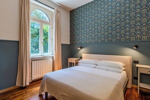 A bedroom with a bed, a window with curtains, a wall with floral wallpaper, and a nightstand with a lamp. - Villa dei Ricordi_Mozzate_Apartment 3 (Mozzate)