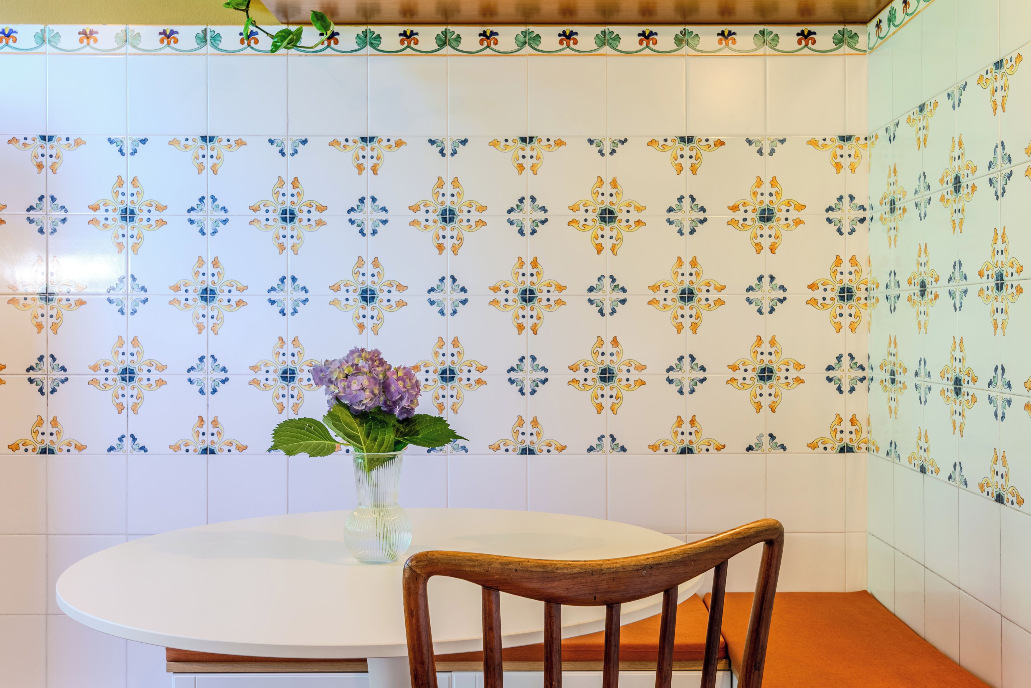 Un'area pranzo con un tavolo rotondo e una sedia di legno, un vaso con fiori e un muro rivestito di piastrelle con motivo floreale.