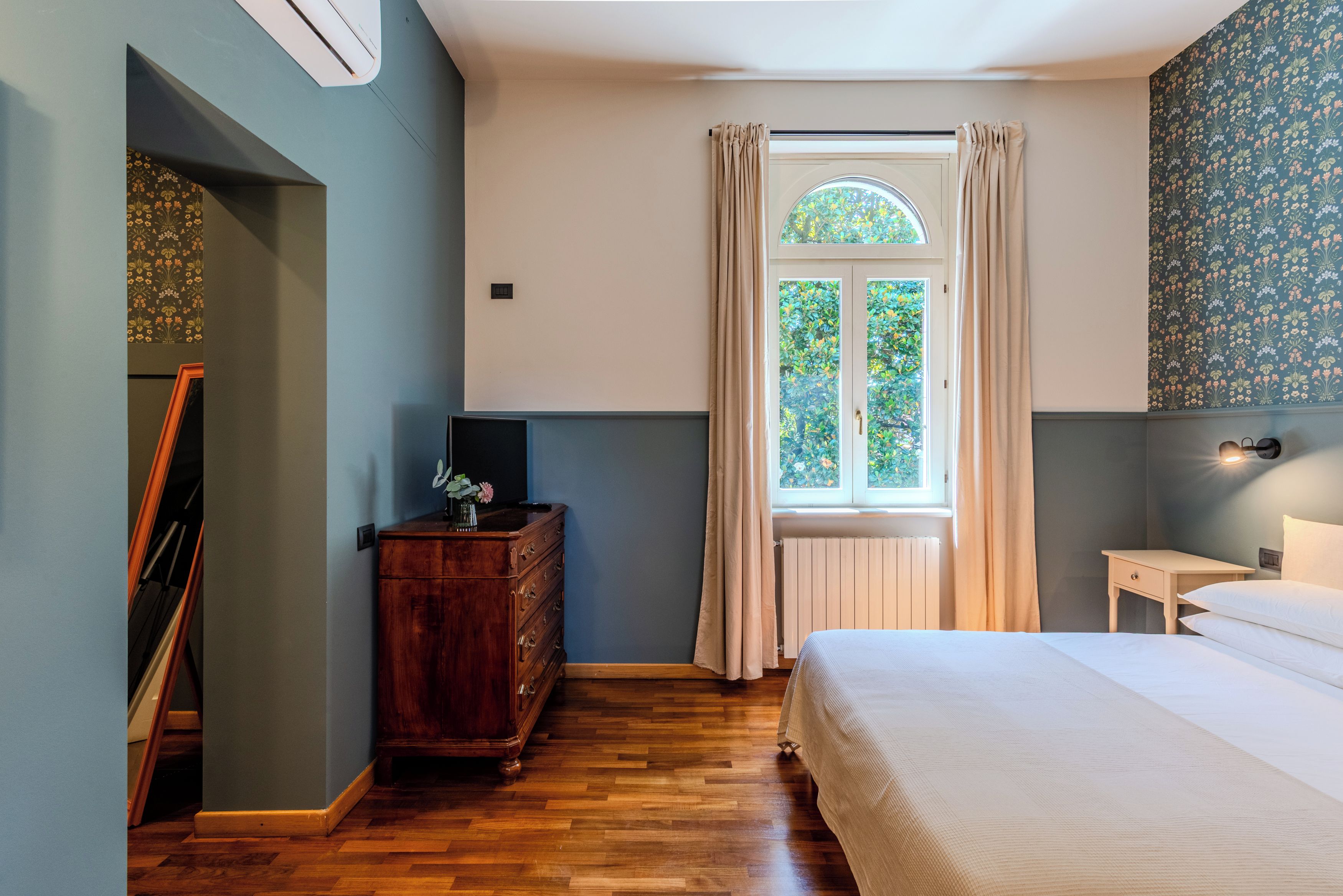 A bedroom with a wooden dresser, a bed, a window with curtains, and a wall with a floral pattern.