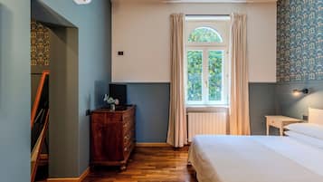A bedroom with a wooden dresser, a bed, a window with curtains, and a wall with a floral pattern.