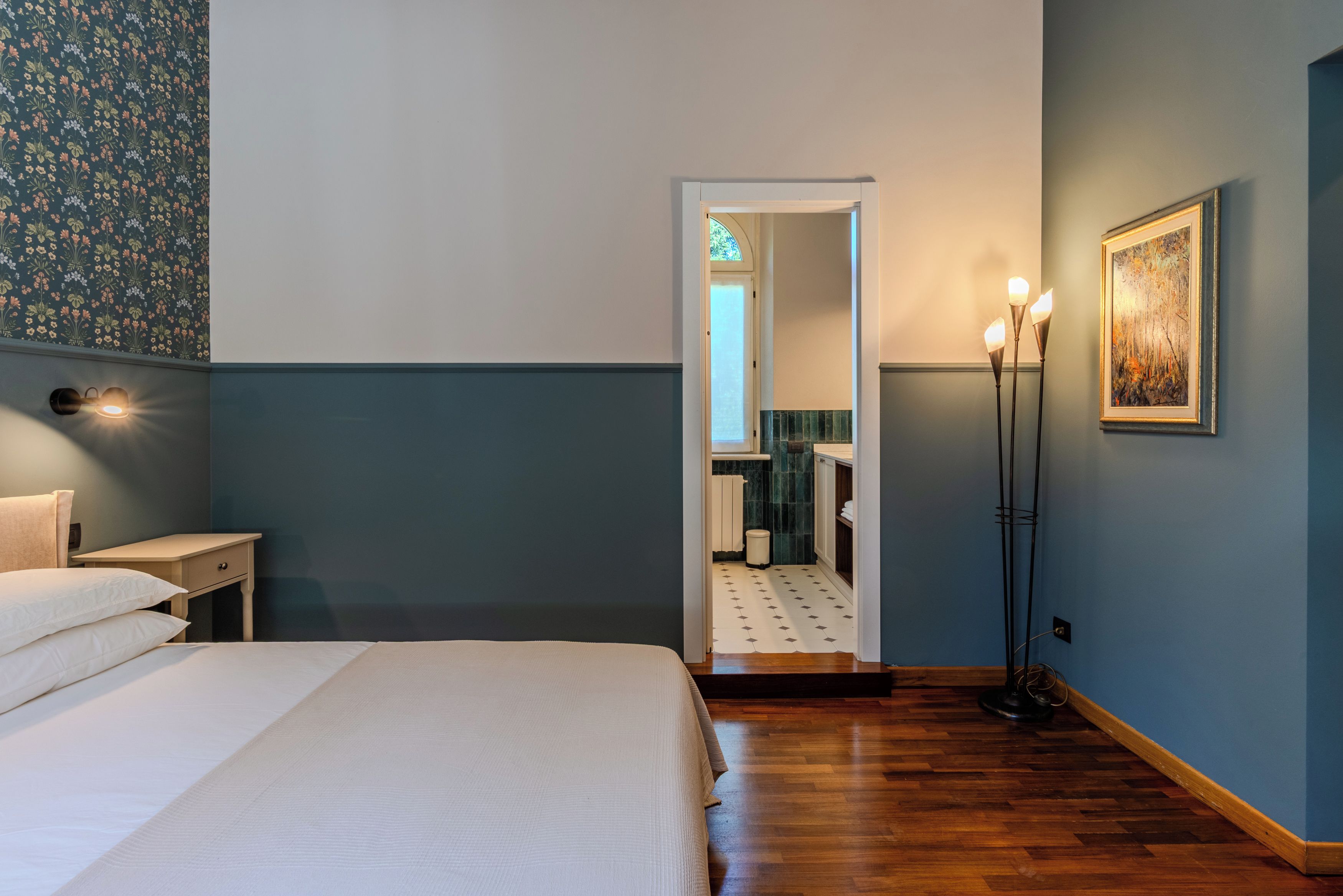 Hotellihuone, jossa on sänky, yöpöytä, seinään kiinnitetty valaisin ja taulu.