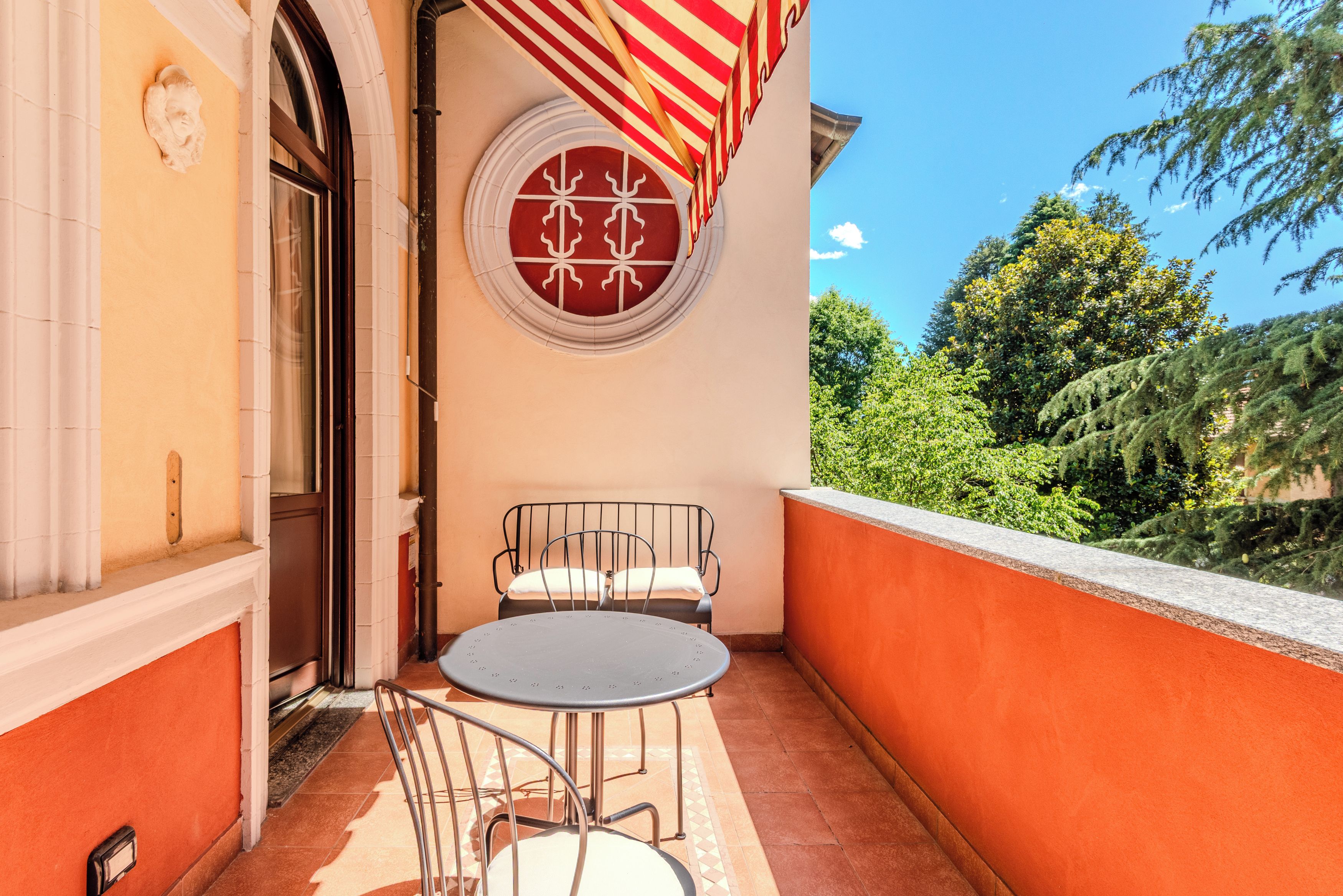 A balcony with a round table and two chairs, a red and white striped awning, and a circular window with a white pattern.