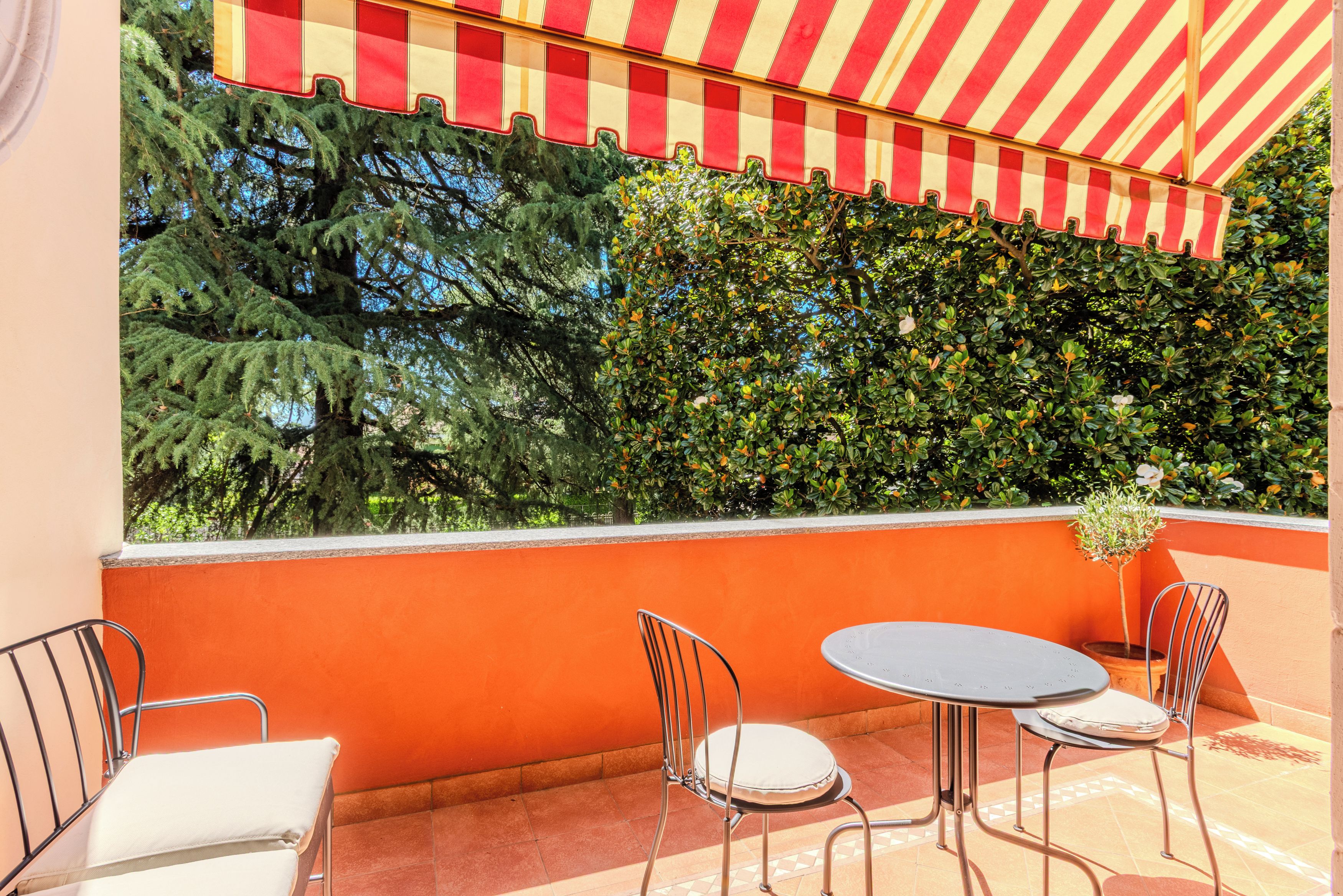 A terrace with a red and white striped awning, two metal chairs, a round table, and a view of green foliage.