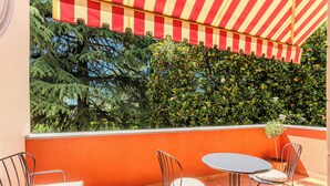 Una terraza con un toldo a rayas rojas y blancas, dos sillas de metal, una mesa redonda y vistas a follaje verde.