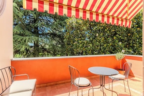 A terrace with a red and white striped awning, two metal chairs, a round table, and a view of green foliage. - Vrbo Property (Mozzate)