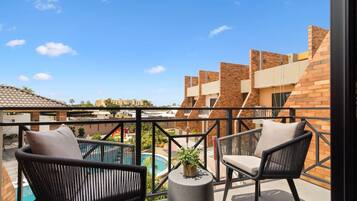 A balcony with two wicker chairs, a small table with a plant, and a view of a swimming pool and buildings.