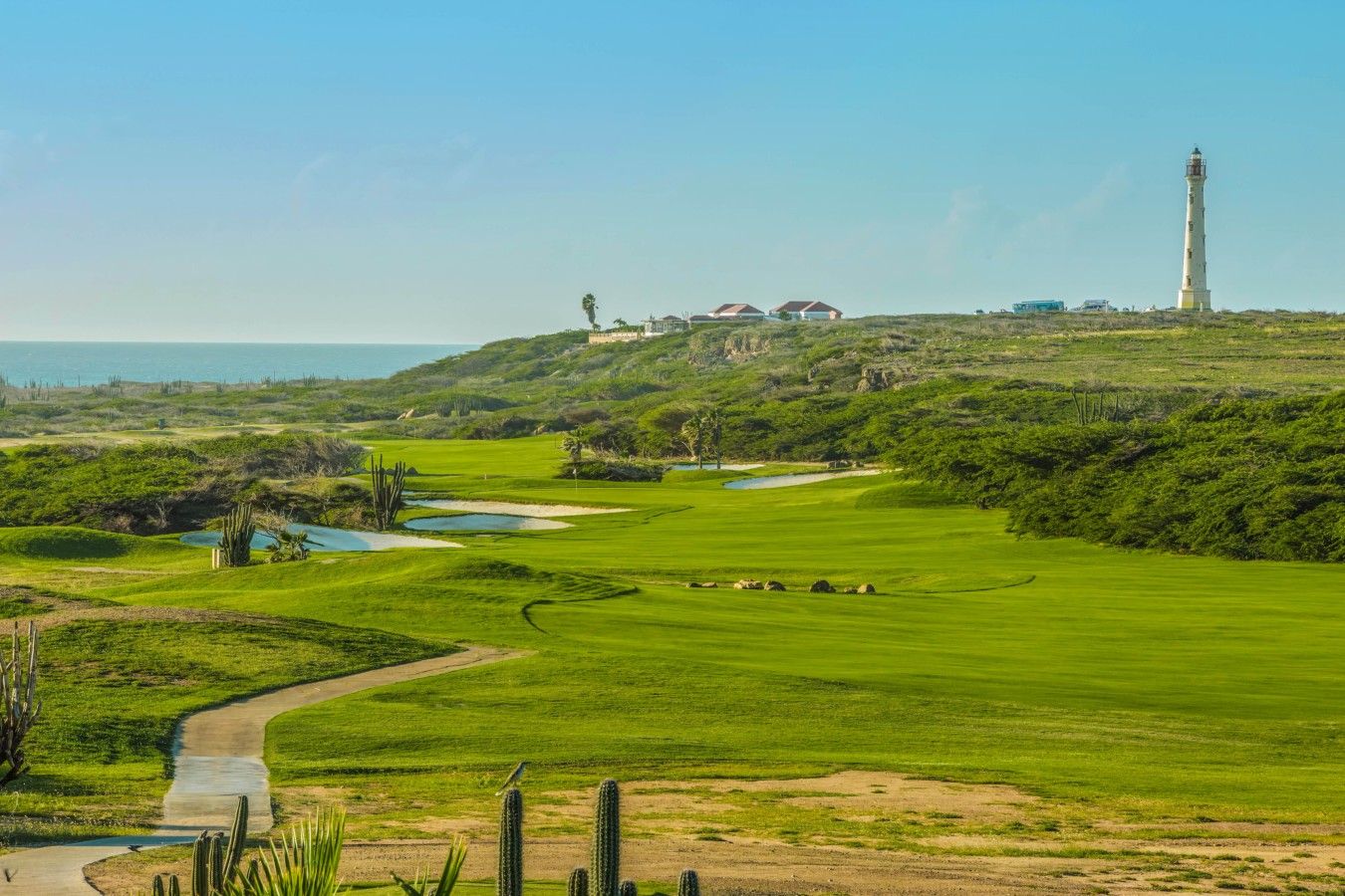 Un campo de golf con búnkeres, un obstáculo acuático, un faro y un edificio en una colina.