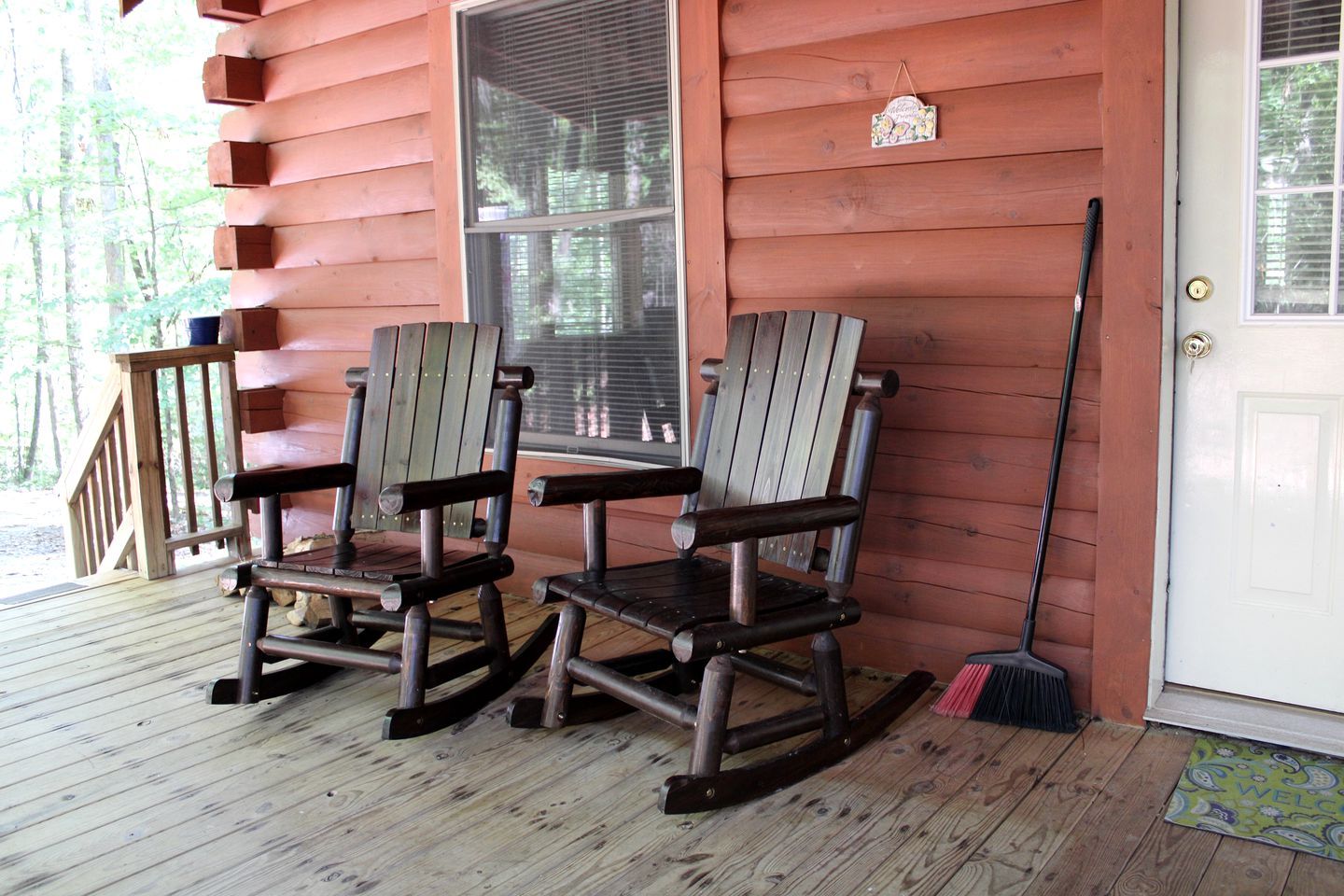 A wooden rocking chair on a porch with a wooden deck, a window, and a door with a welcome mat.