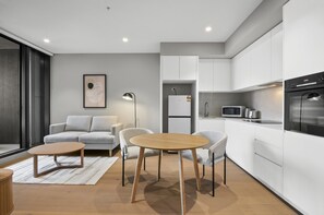 A modern apartment with a living area featuring a sofa, two armchairs, a coffee table, and a floor lamp. The kitchen includes white cabinets, a microwave, and a refrigerator. - Skyline 1-bed Apartment Near Melbourne CBD (Melbourne)