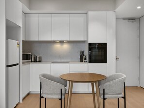 A modern kitchen with white cabinetry, a black oven, a microwave, and a coffee maker. There is a round wooden table with two grey upholstered chairs. - Skyline 1-bed Apartment Near Melbourne CBD (Melbourne)