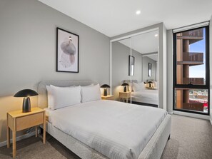 A modern bedroom with a large bed, bedside tables, a mirror, and a city view. - Skyline 1-bed Apartment Near Melbourne CBD (Melbourne)
