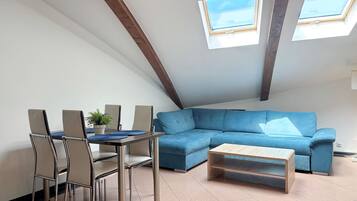 Une chambre de style loft moderne avec un plafond en pente, des fenêtres de toit, un canapé d’angle bleu, une table basse en bois et un coin repas avec des chaises et une petite plante.