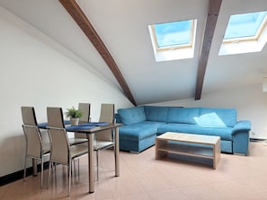 A modern loft bedroom with a sloped ceiling, skylights, a blue sectional sofa, a wooden coffee table, and a dining area with chairs and a small plant. - MARGI City 14 Apartment (Szczecin)