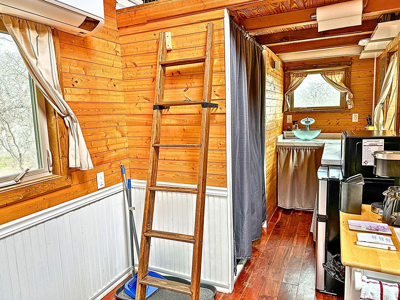 Interior de una cabaña de madera con una escalera, una ventana, un lavamanos y un refrigerador.