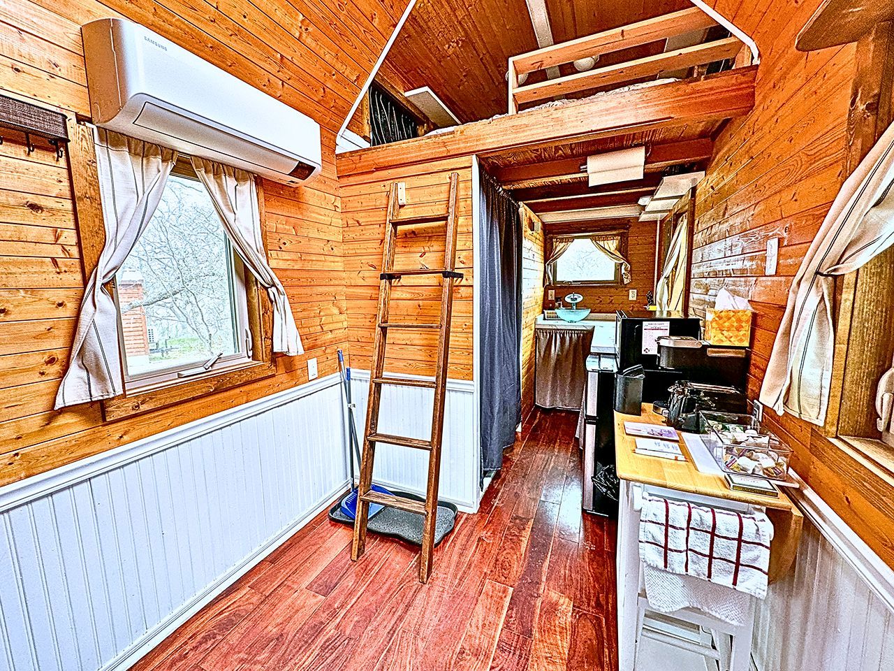 A wooden interior with a kitchen area, a ladder, and a window with curtains.