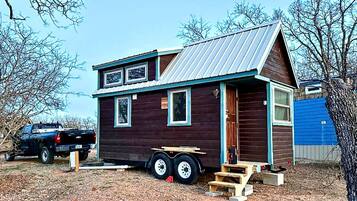 A small, two-story cabin with a metal roof, a front porch, and a side entrance.