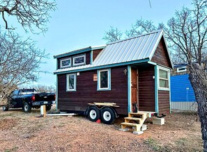 A small, two-story cabin with a metal roof, a front porch, and a side entrance.