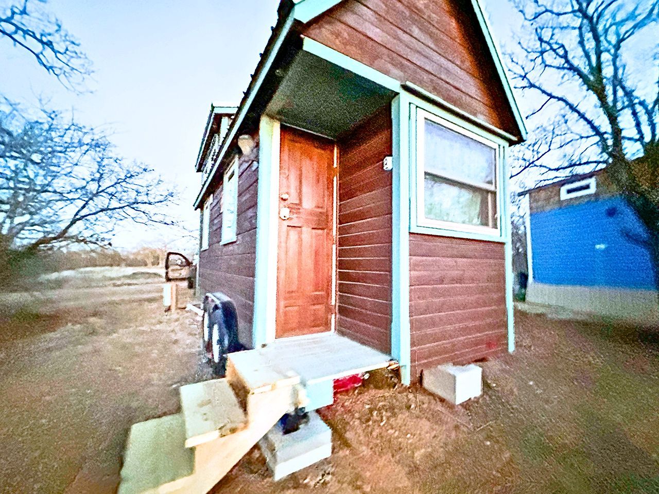 A small wooden cabin with a single door and a window, a step leading up to the door, and a blue garage door in the background.
