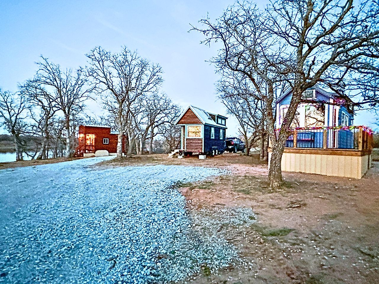 A rural property with a gravel driveway, a small wooden cabin, and a larger house with a fenced yard.