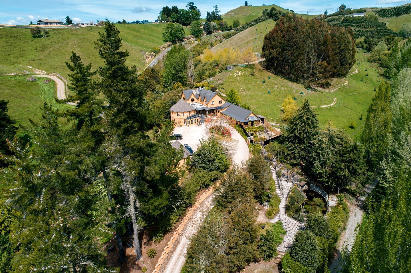 A large house with a spacious driveway and a well-maintained lawn, surrounded by dense trees and rolling hills.