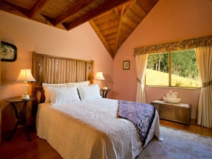 A bedroom with a wooden ceiling, a bed with white bedding, two bedside tables with lamps, a window with curtains, and a small table with a decorative item.