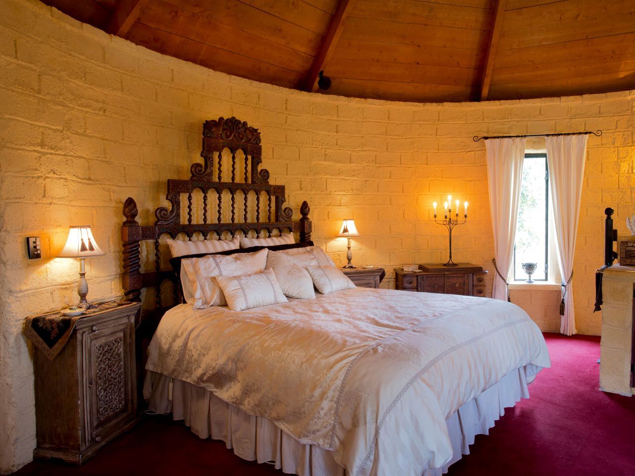 A bedroom with a large bed, wooden headboard, two bedside tables with lamps, a window with curtains, and a brick wall.