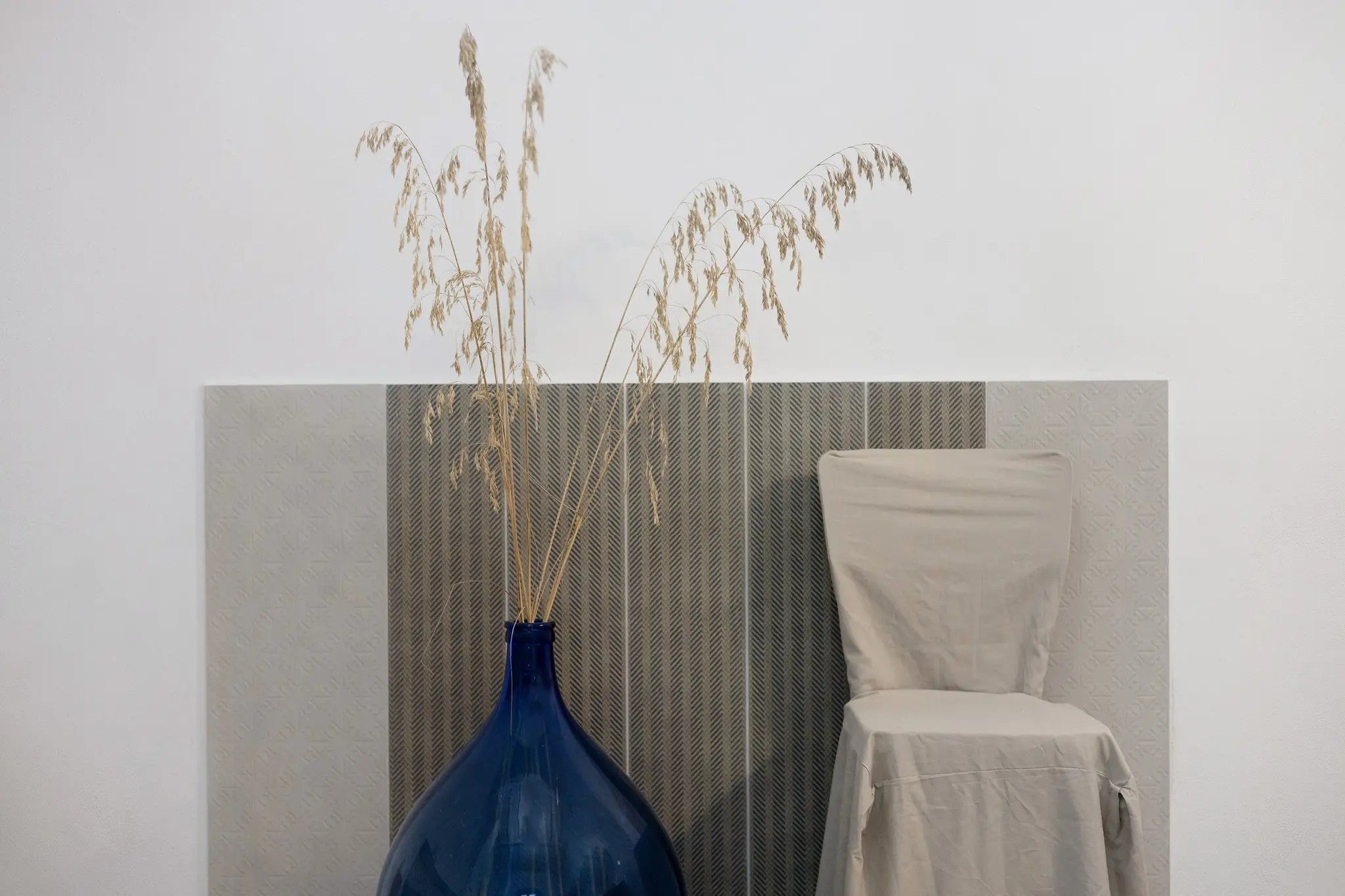 A room with a blue vase containing dried plants, a chair, and a patterned wall.