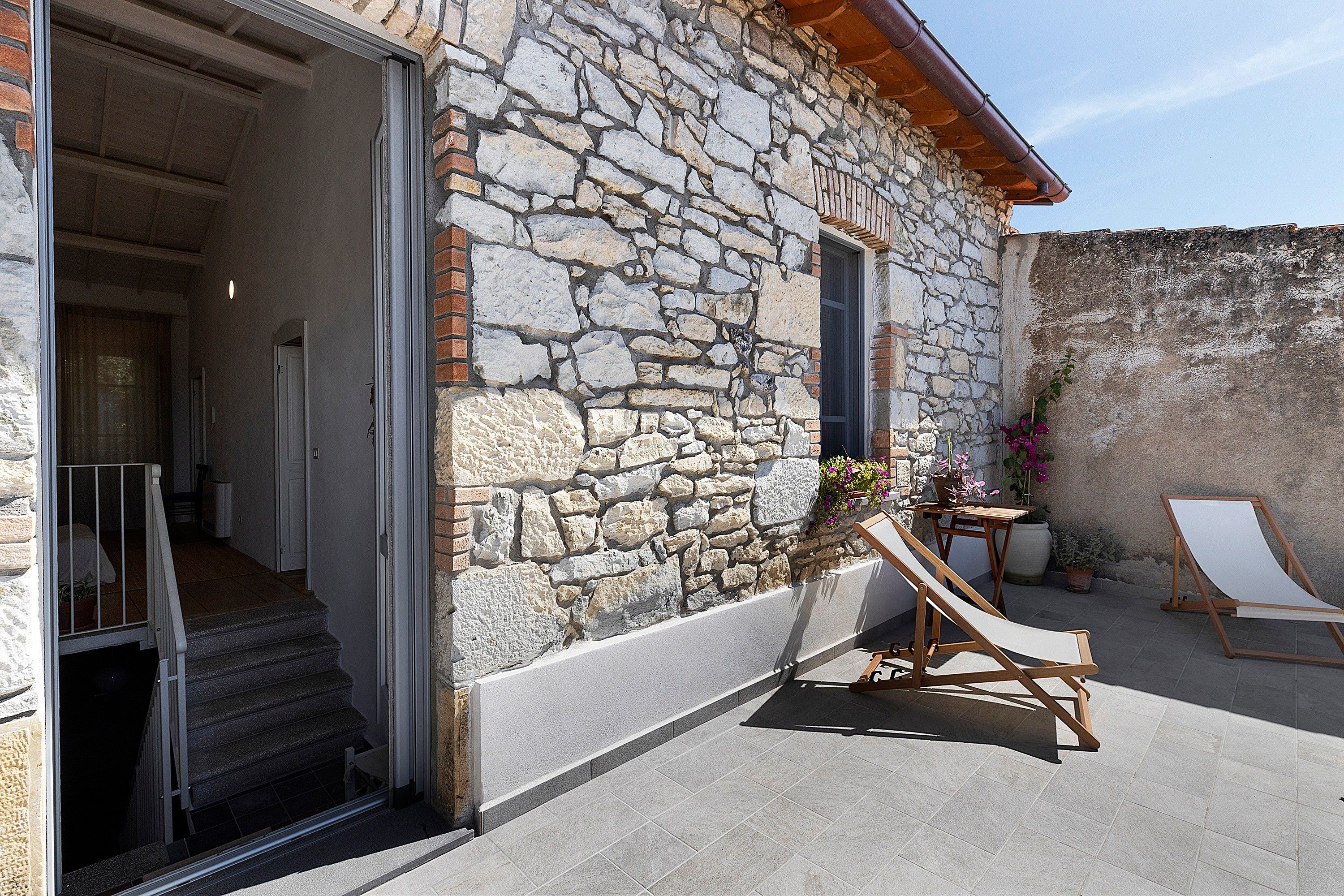 Un bâtiment en pierre avec un escalier en bois menant à une porte ouverte, une petite table ornée de plantes en pot et un fauteuil de jardin en bois.