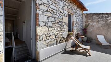 Un bâtiment en pierre avec un escalier en bois menant à une porte ouverte, une petite table ornée de plantes en pot et un fauteuil de jardin en bois.