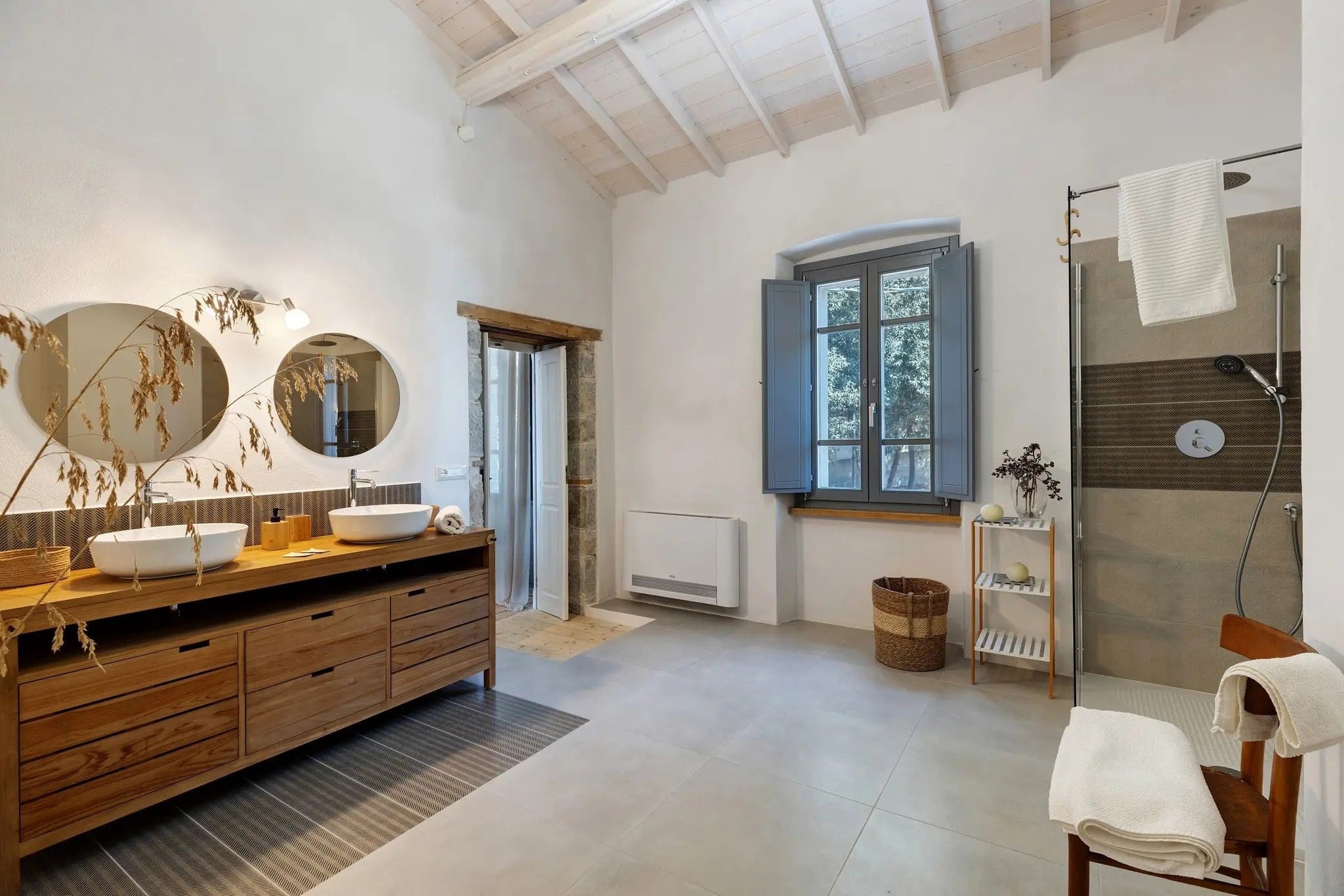 Un bagno moderno con un mobile vanitario in legno, uno specchio rotondo, un'ampia finestra e uno spazio doccia.