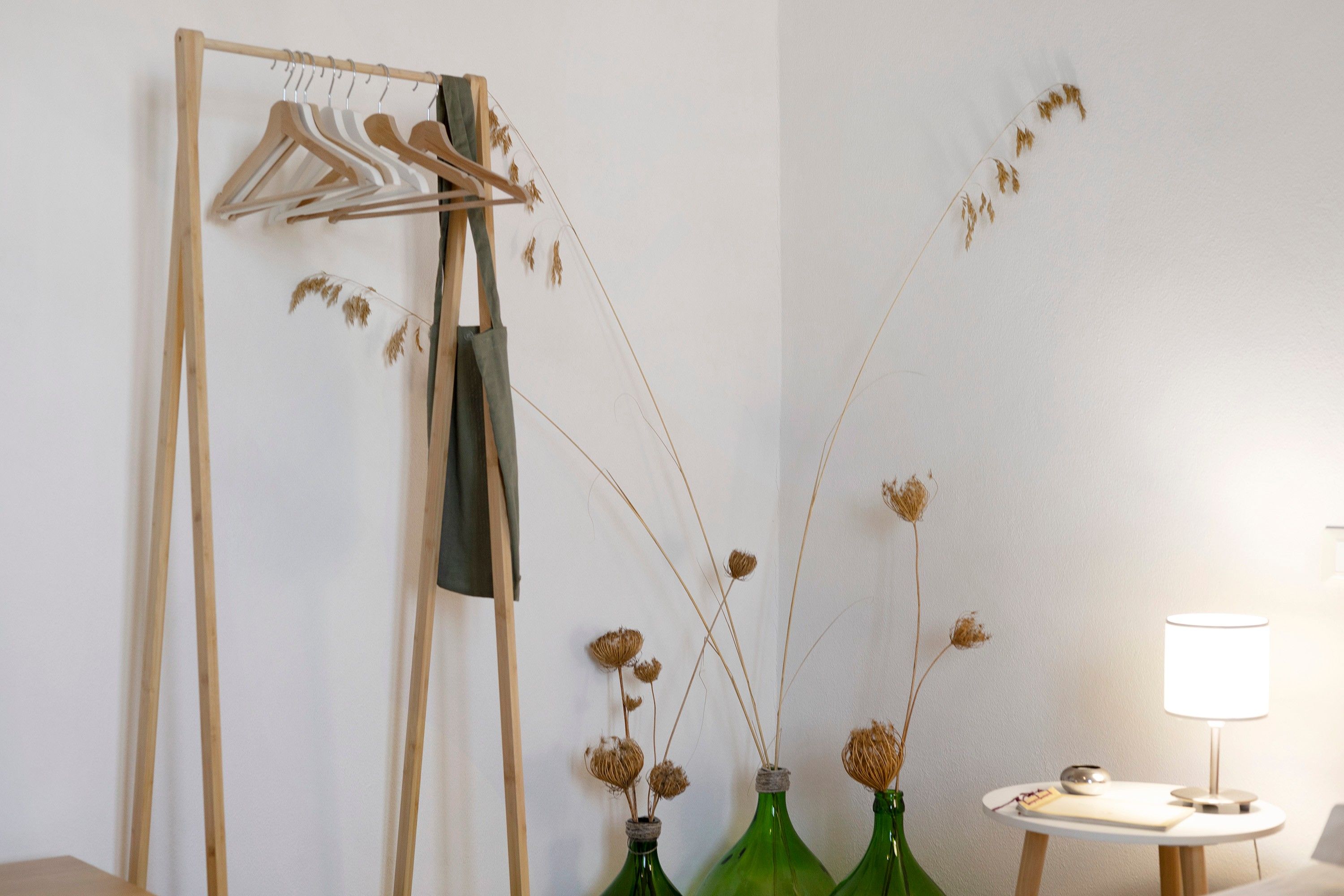 Une pièce avec un porte-manteau en bois, trois bouteilles vertes contenant des plantes séchées, une petite table ronde avec une lampe et un mur blanc.