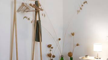 Une pièce avec un porte-manteau en bois, trois bouteilles vertes contenant des plantes séchées, une petite table ronde avec une lampe et un mur blanc.