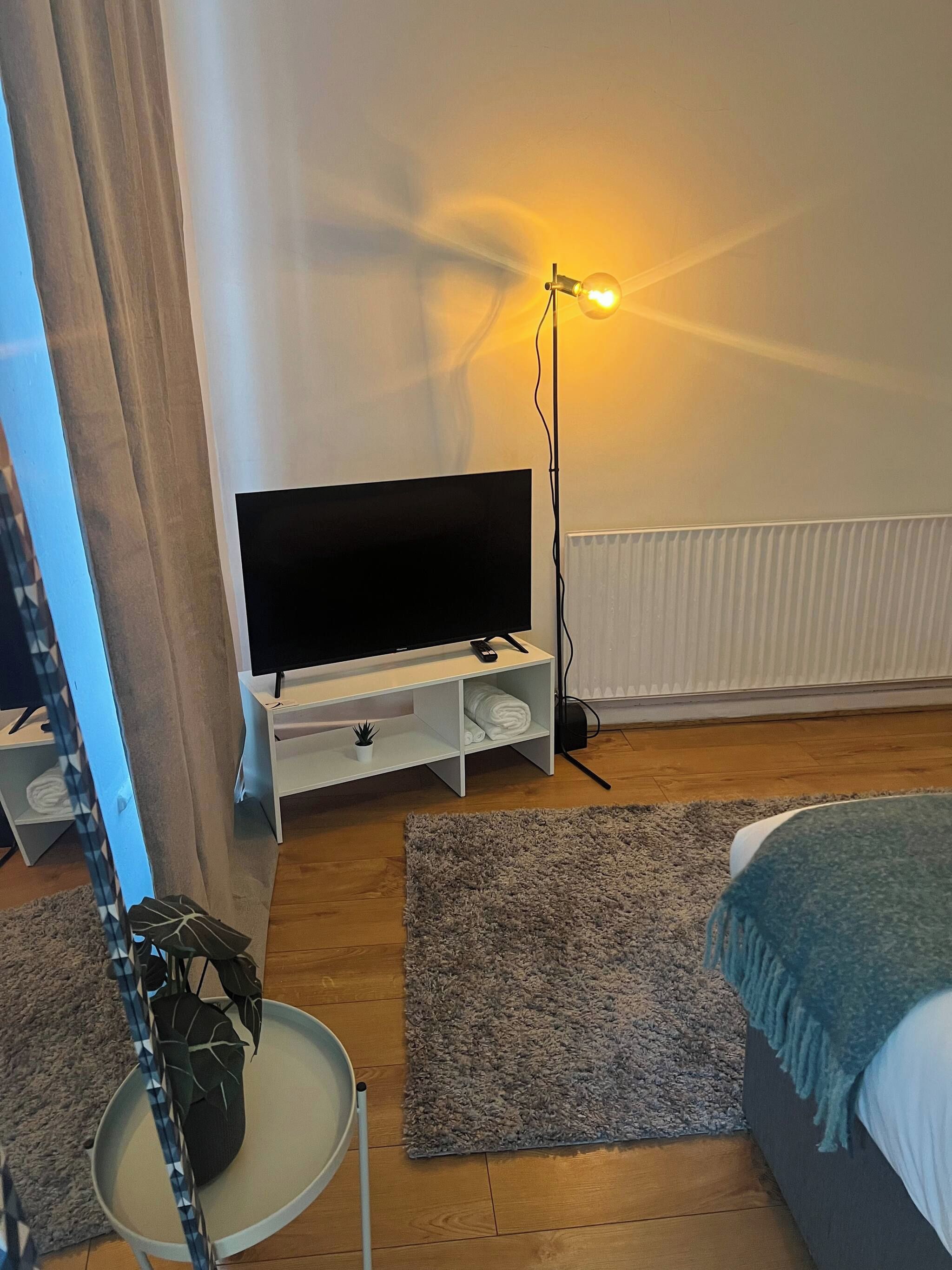 Une chambre à coucher avec un téléviseur posé sur un meuble, une table de chevet, une lampe sur pied, un lit, un tapis gris et une fenêtre avec des rideaux.