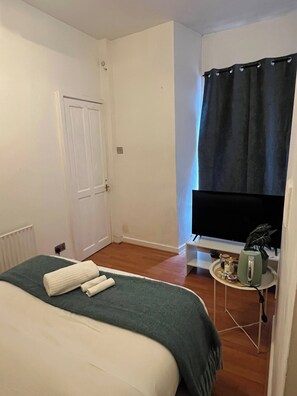 Ein Schlafzimmer mit einem Bett, einem Fernseher, einem kleinen Tisch mit Wasserkocher und einer Tür.