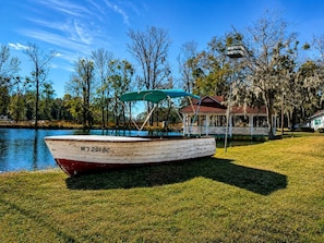 Un bateau portant le numéro « M7 201 BC » est amarré sur une rive herbeuse au bord d’un lac. On aperçoit, en arrière-plan, un kiosque et une maison.