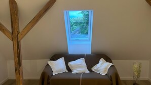 Ein Zimmer mit einem braunen Sofa, drei weißen Kissen, einem Holzbalkenbau, einem Oberlicht und einer Vase mit Blumen.