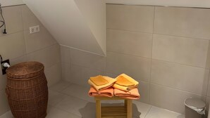 Ein kleines Badezimmer mit schräg versiegelter Decke, einem Holzschemel mit gefalteten Handtüchern, einem Korbwäsches und einem Mülleimer.