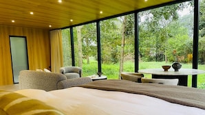 Une chambre moderne dotée d’un grand lit, d’un coin salon avec des fauteuils, d’une petite table et offrant une vue sur un jardin luxuriant grâce à de grandes fenêtres.