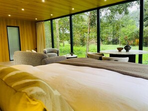 A modern bedroom with a large bed, a seating area with chairs, a small table, and a view of a lush garden through large windows. - S.P.A. Parel in 't Groen (Moergestel)